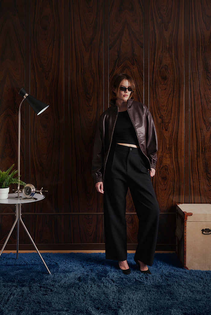 A woman stands in a room with dark wood paneling and a blue rug, wearing a black crop top, black wide-leg pants, and a dark brown leather jacket. 