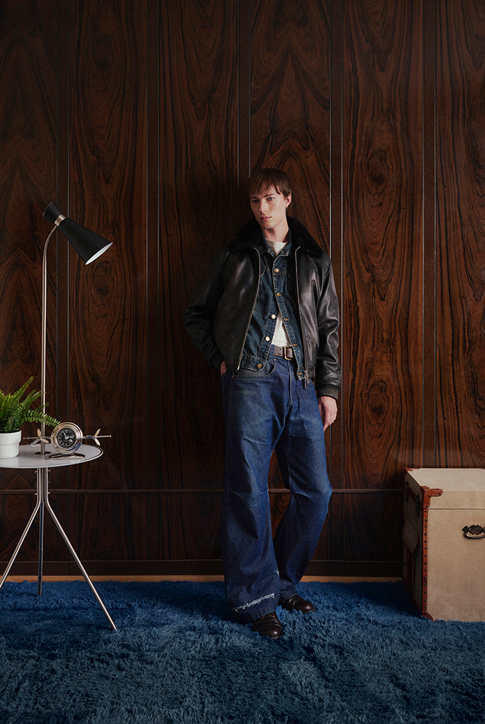 A model stands in a room with dark wood-paneled walls and a blue shag rug, wearing a black leather jacket distress finishing from Sly&Co