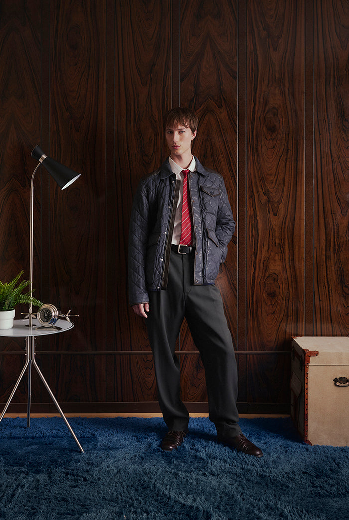 A man stands in a room with dark wood-paneled walls and a blue shag rug, wearing a navy quilted leather jacket from Sy&Co