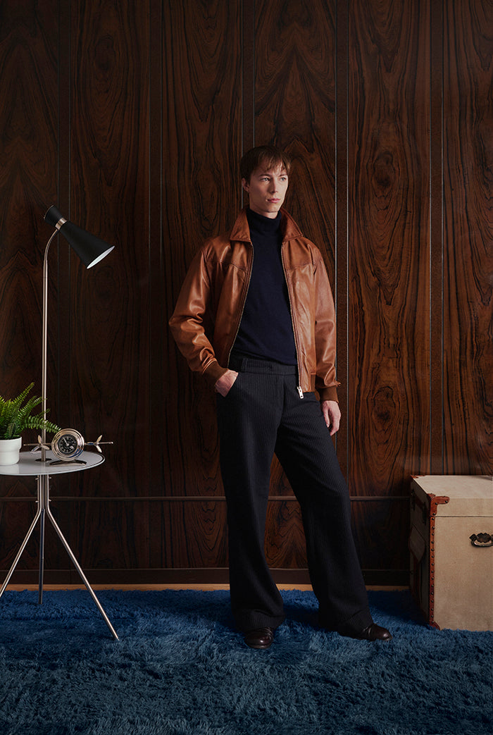 A male model wearing a caramel color leather jacket with a front zipper, pointed collar, and ribbed knit cuffs and hem. City-Sly&Co