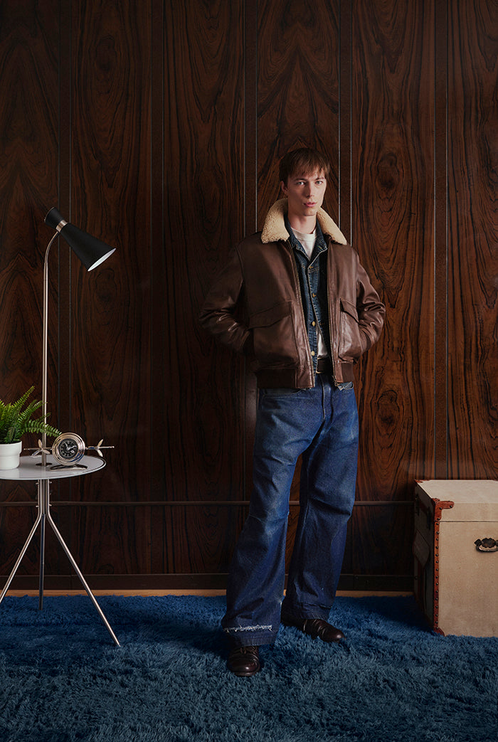 A man stands in a room wearing a brown leather aviator-style jacket. D Four-Dly&Co