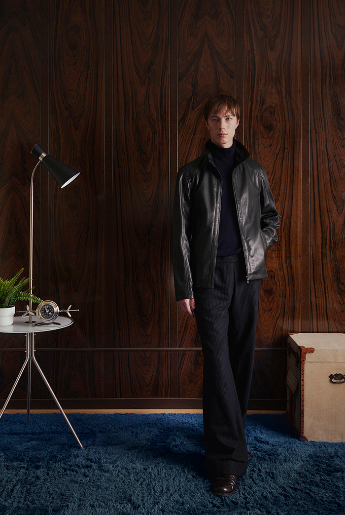 A model stands in a room with dark wood-paneled walls and a blue rug, wearing a black leather jacket, Dolci from Sly&Co