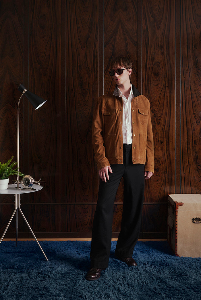 A person stands in a styled interior wearing a brown suede jacket over a white shirt and black trousers.