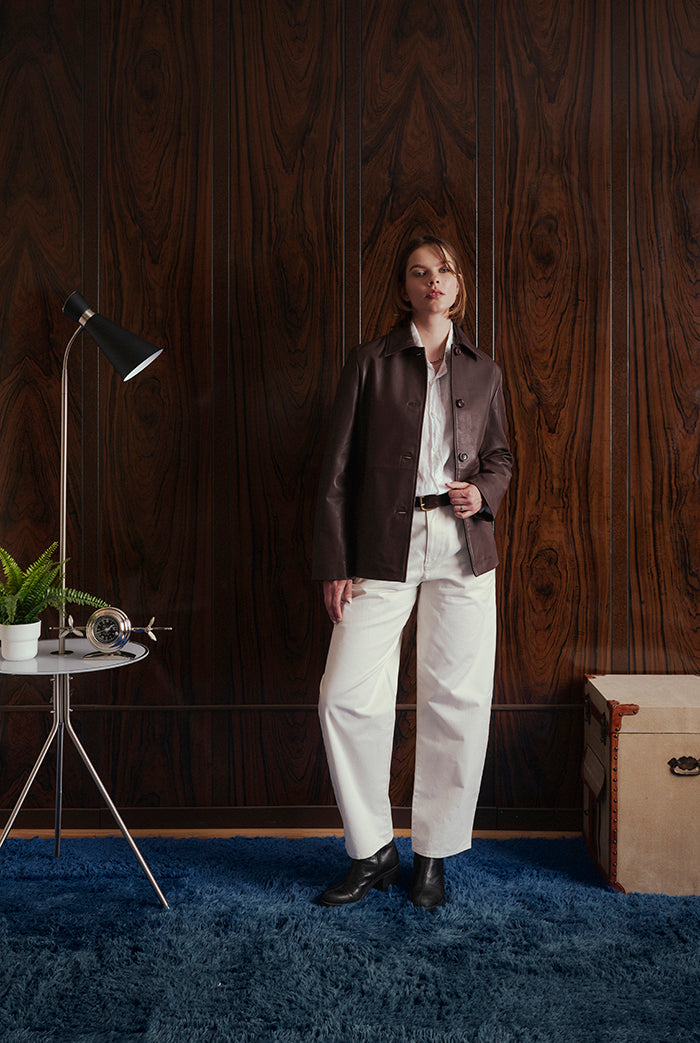 A model stands in a room with dark wood-paneled walls, wearing a dark brown leather jacket, white shirt, and white trousers.