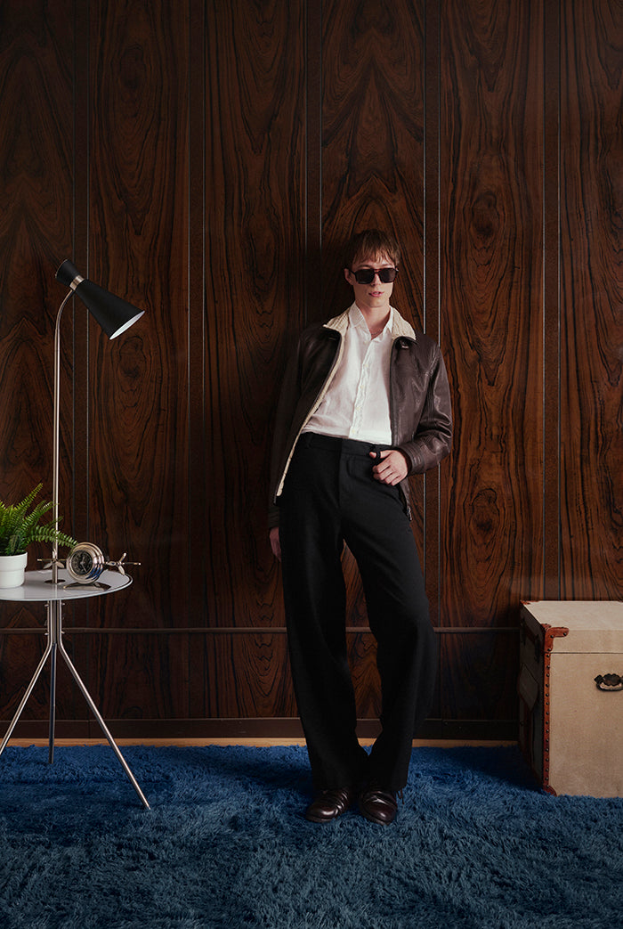 A person stands in a styled interior wearing a brown leather jacket, white collared shirt, black trousers, and dark sunglasses, posed against a dark wood-paneled wall with a blue rug.