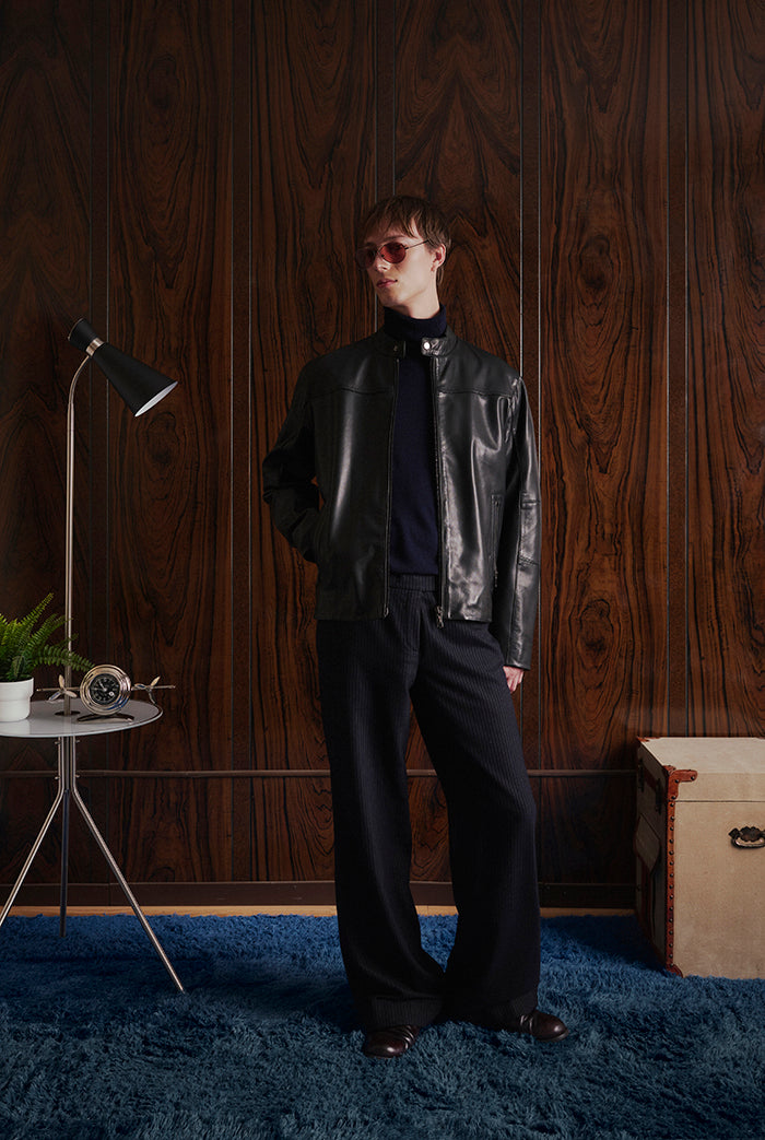 A model stands in a room with dark wood-paneled walls wearing a black leather jacket from Sly&Co