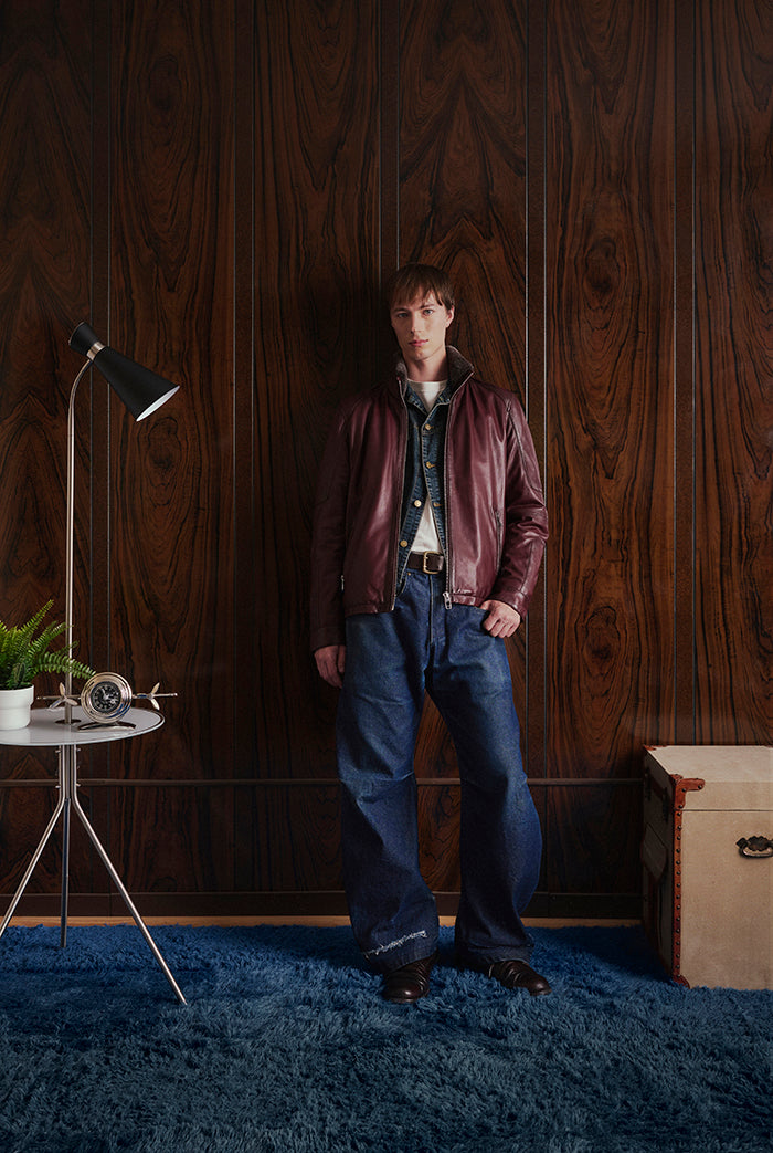 A man stands in a room with dark wood-paneled walls and a blue shag rug, wearing a burgundy leather jacket.