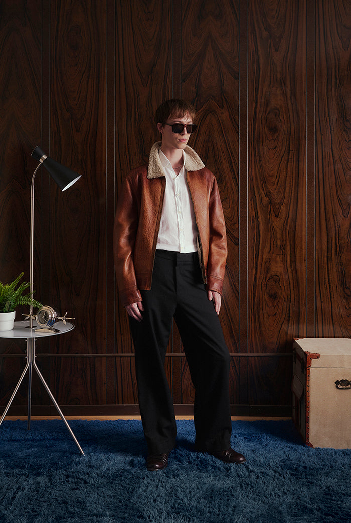 A model stands in a room wearing a brown leather jacket from Sly&Co