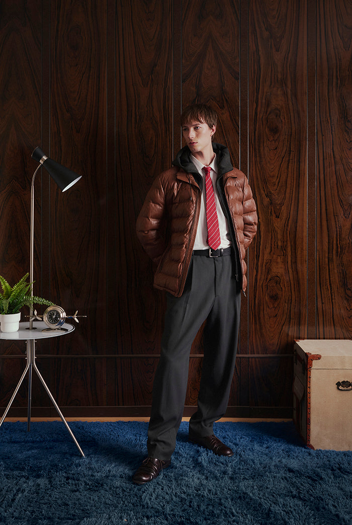 A male model stands in a room wwearing a brown puffer leather jacket with an attached hoodie. Ryley_ Sly&Co