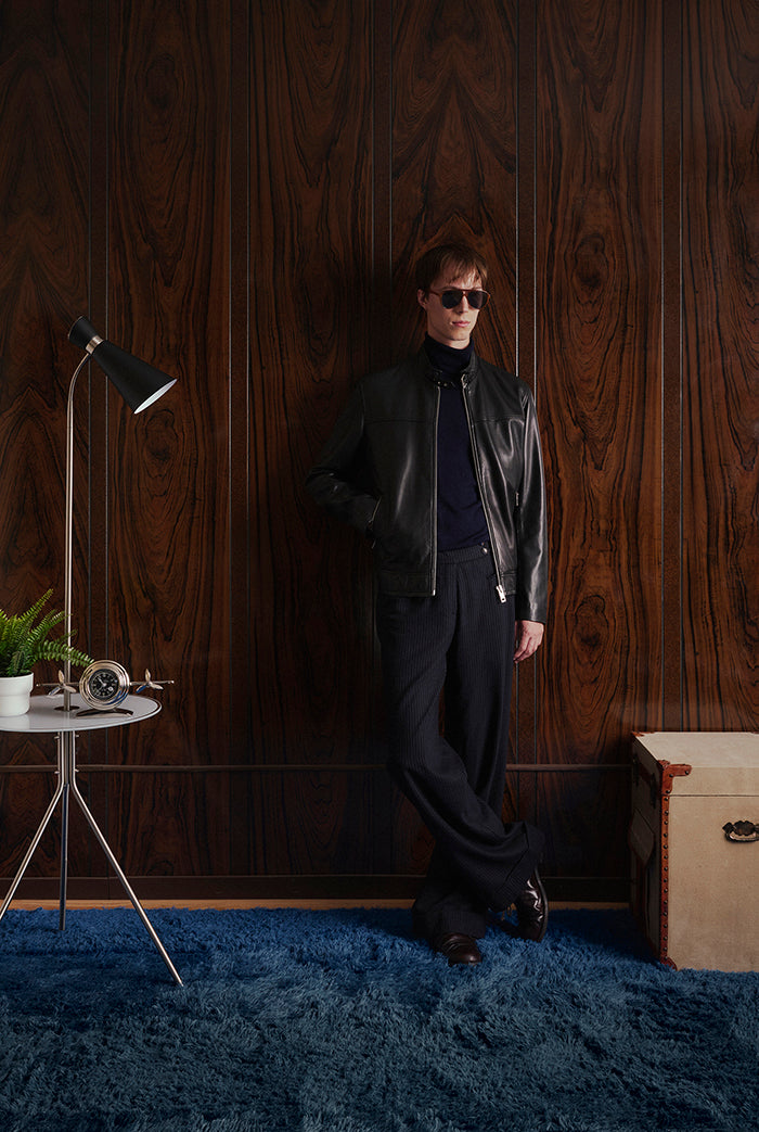 A male model wearing a black leather jacket with a front zipper, stand collar, and two zippered hand pockets. Vespa from Sly&Co